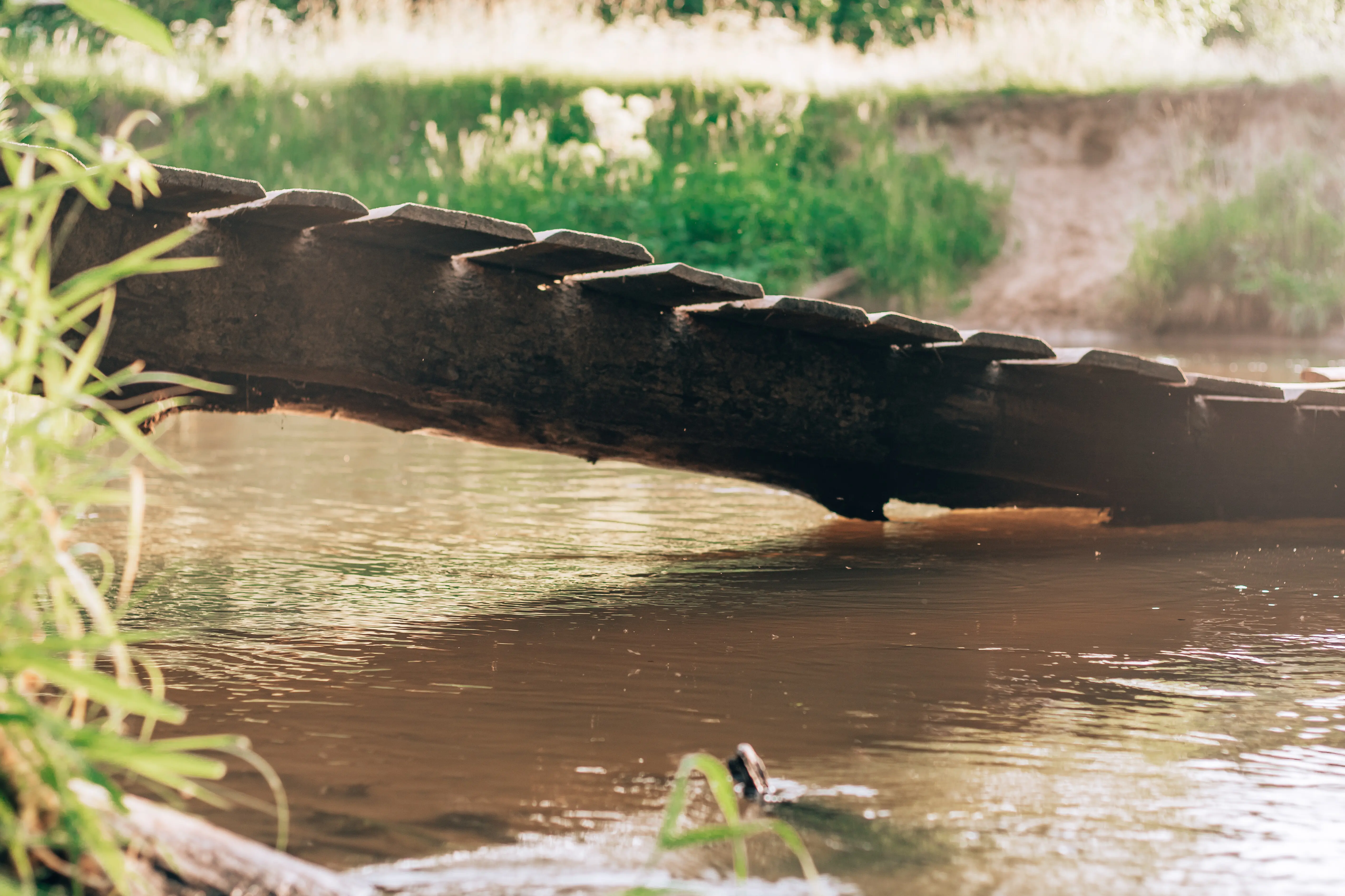 A tilted, crooked wooden bridge crossing a small river, constructed from old planks.